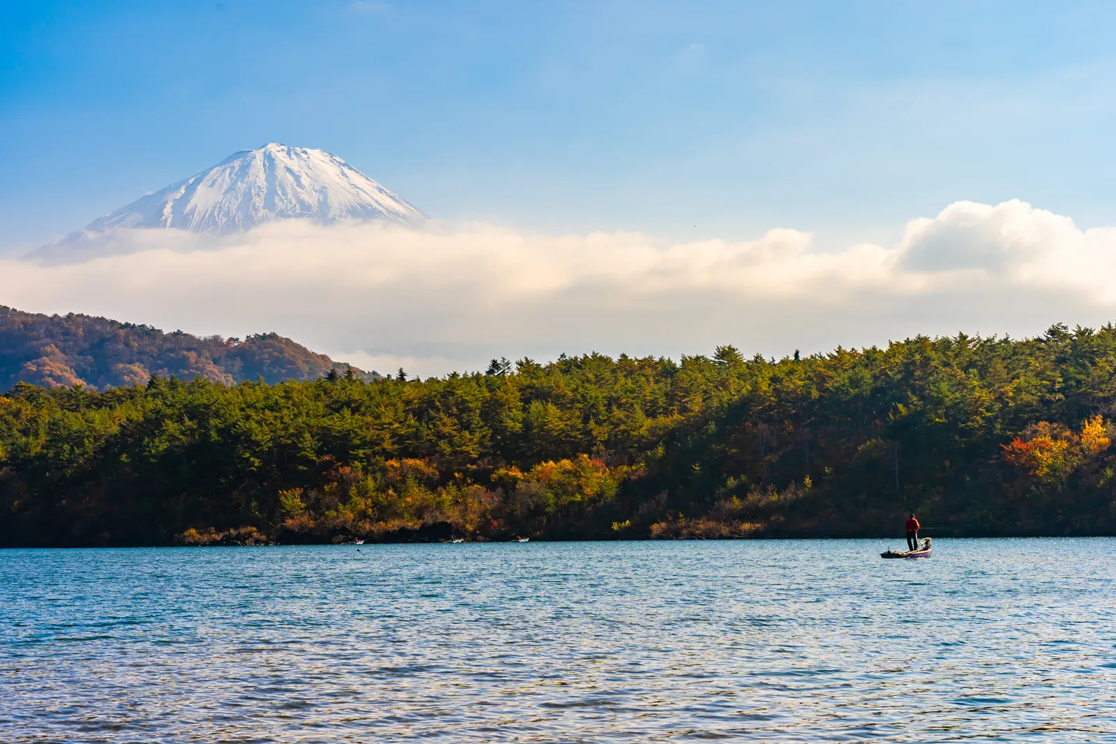 The snow-capped cone of Mount Fuji rising above the clouds over a lake and forests